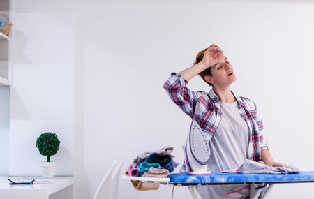 beautiful young red haired housewife in good mood ironing clothes on ironing board in front of a white wall at homeの写真素材
