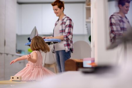 happy family having fun together at home  cute little daughter in a pink dress playing and dancing while young redhead mother ironing clothes behind herの写真素材