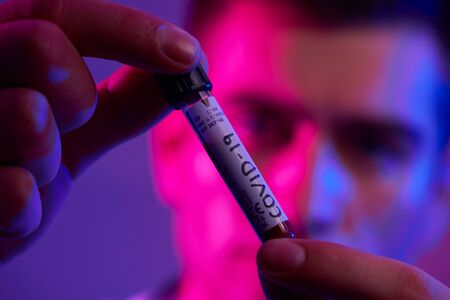 Coronavirus, Doctor holding positive covid-19 virus Blood Sample test tube. Wearing biohazard epidemic Protective mask, suit and glows neon light backgroundの写真素材