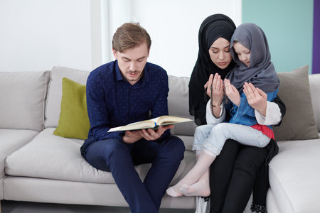 Traditional muslim family parents with children reading Quran and praying together on the sofa...