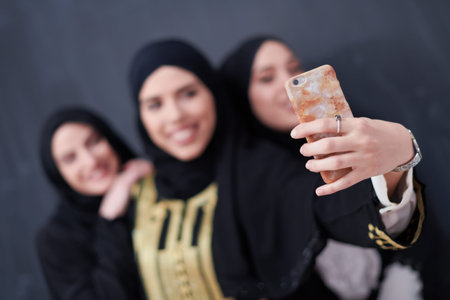 group of young beautiful muslim women in fashionable dress with hijab using mobile phone while taking selfie picture in front of black chalkboard representing modern islam fashion technology and ramadan kareem conceptの写真素材