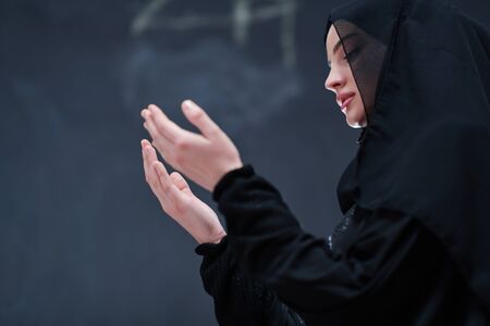 portrait of beautiful muslim woman in fashionable dress with hijab making traditional prayer to God keeps hands in praying gesture isolated on black chalkboard background representing modern islam fashion and ramadan kareem conceptの写真素材
