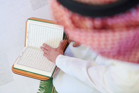 young arabian muslim man in traditional clothes reading holy book Quran on the praying carpet before iftar dinner during a ramadan feast at homeの写真素材