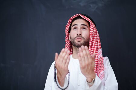 young arabian man in traditional clothes making traditional prayer to God, keeps hands in praying gesture in front of black chalkboard representing modern islam fashion and ramadan kareem conceptの写真素材