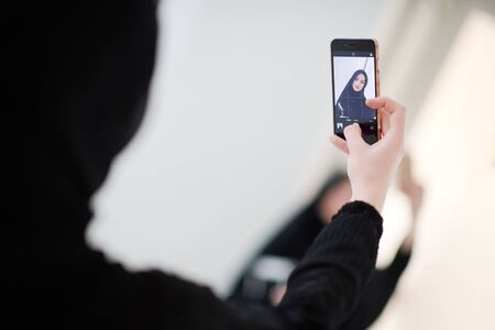 young beautiful muslim woman in fashionable dress with hijab using mobile phone while taking selfie picture at home representing modern islam fashion technology and ramadan kareem conceptの写真素材