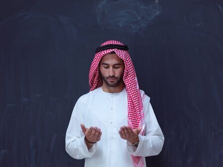 young arabian man in traditional clothes making traditional prayer to God, keeps hands in praying gesture in front of black chalkboard representing modern islam fashion and ramadan kareem conceptの写真素材