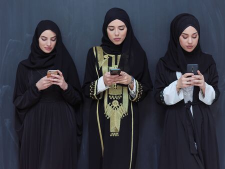 group of beautiful muslim women in fashionable dress with hijab using mobile phone in front of black chalkboard representing modern islam fashion technology and ramadan kareem conceptの写真素材
