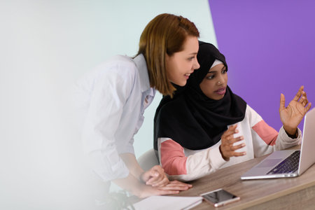 afro muslim woman wearing a hijab sits smiling in her home office and using laptop for online...