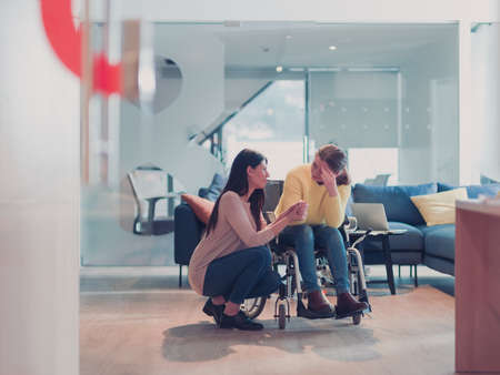 Sad and depressed disabled businesswoman in a wheelchair at the office getting support and assistance of female coworker friendの写真素材