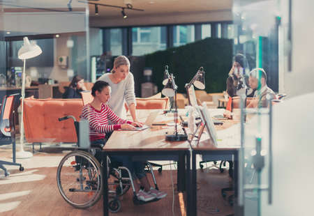 Disabled businesswoman in a wheelchair in modern coworking office space. Colleagues in background. Disability and handicap concept. Selective focusの写真素材