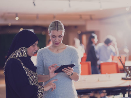 Blonde and modern Muslim businesswoman friends collaborating as a team and working on tablet. Diverse team in modern open space office in backgroundの写真素材