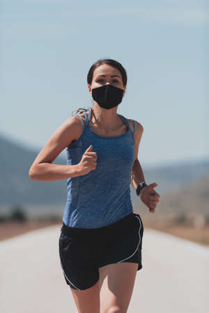 Determined fitness woman in short clothes wearing red protective face mask running outdoors in the city during coronavirus outbreak. Covid 19 and physical jogging activity, sport and fitness.の写真素材