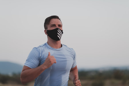 Fitness man in wet sporty clothes wearing black protective face mask running outdoors in the city during coronavirus outbreak. Covid 19 and physical jogging activity sport and fitness. New normalの写真素材