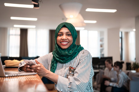 African Muslim business woman wearing a green hijab using smartphone while working on laptop...