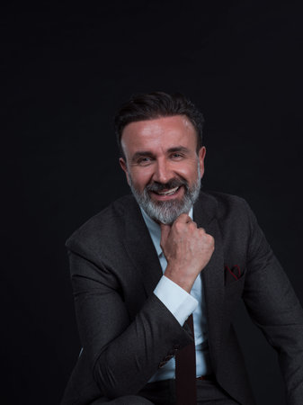 Portrait of adult businessman wearing trendy suit and sitting in modern studio on stylish chair against the black backgroundの写真素材