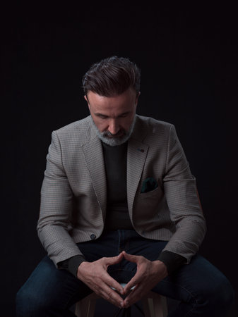 Portrait of adult businessman wearing trendy suit and sitting in modern studio on stylish chair against the black backgroundの写真素材