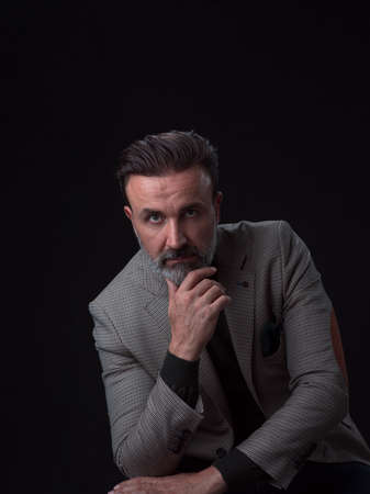 Portrait of adult businessman wearing trendy suit and sitting in modern studio on stylish chair against the black backgroundの写真素材