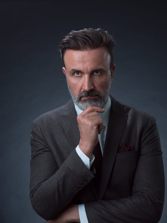 Portrait of a stylish elegant senior businessman with a beard and casual business clothes in photo studio isolated on dark background gesturing with handsの写真素材