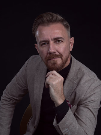 Portrait of adult businessman wearing trendy suit and sitting in modern studio on stylish chair...