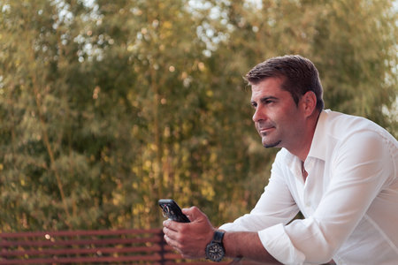 A businessman in casual clothes resting on the balcony of the terrace while using a smartphone....