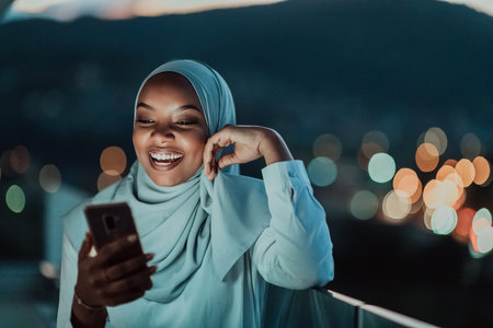 Young Muslim woman wearing scarf veil on urban city street at night texting on a smartphone with...