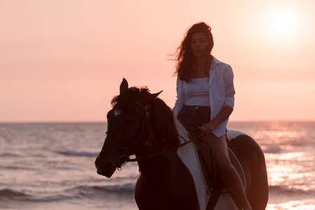Woman in summer clothes enjoys riding a horse on a beautiful sandy beach at sunset. Selective focusの写真素材