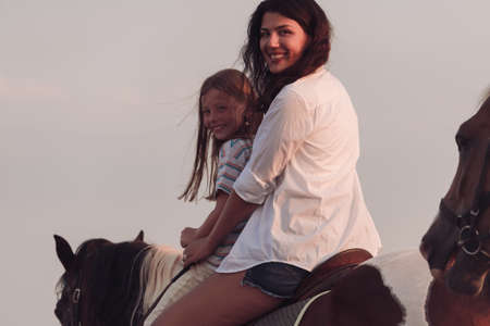 The family spends time with their children while riding horses together on a beautiful sandy beach on sunet.の写真素材