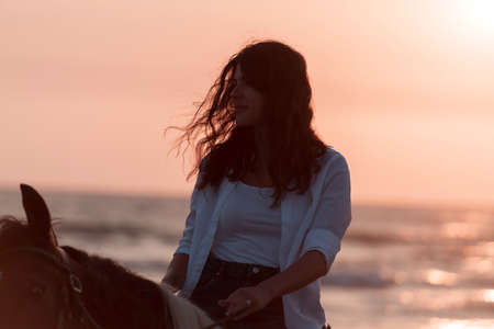 Woman in summer clothes enjoys riding a horse on a beautiful sandy beach at sunset. Selective focusの写真素材
