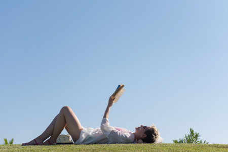 Woman lying and reading her favorite book on a meadow covered with fresh green grass on a sunny...
