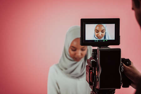 Videographer in digital studio recording video on professional camera by shooting female Muslim woman wearing hijab scarf plastic pink background.の写真素材
