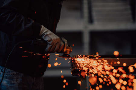 Heavy Industry Engineering Factory Interior with Industrial Worker Using Angle Grinder and Cutting a Metal Tube. Contractor in Safety Uniform and Hard Hat Manufacturing Metal Structures.の写真素材