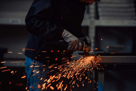 Heavy Industry Engineering Factory Interior with Industrial Worker Using Angle Grinder and Cutting a Metal Tube. Contractor in Safety Uniform and Hard Hat Manufacturing Metal Structures.の写真素材