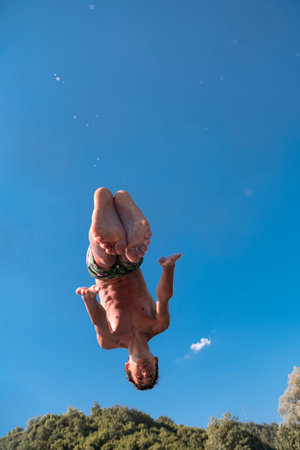 Young teen boy jumping flying and diving in the river. Clear blue sky and trees in distance as a...