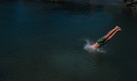 Young teen boy jumping flying and diving in the river. Clear blue sky and trees in distance as a...