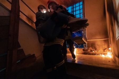 Brave Fireman Descends Stairs of a Burning Building and Holds Saved Girl in His Arms. Open fire and one Firefighter in the Background.の写真素材