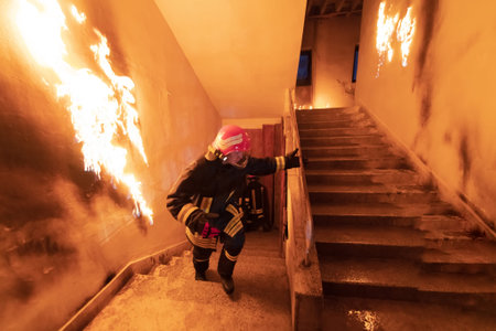 Brave Fireman Descends Stairs of a Burning Building and Holds Saved Girl in His Arms. Open fire and one Firefighter in the Background.の写真素材