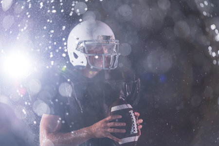 American Football Field: Lonely Athlete Warrior Standing on a Field Holds his Helmet and Ready to Play. Player Preparing to Run, Attack and Score Touchdown. Rainy Night with Dramatic Fog, Blue Lightの写真素材