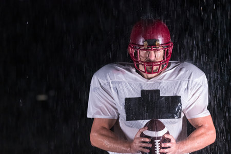American Football Field: Lonely Athlete Warrior Standing on a Field Holds his Helmet and Ready to Play. Player Preparing to Run, Attack and Score Touchdown. Rainy Night with Dramatic Fog, Blue Lightの写真素材
