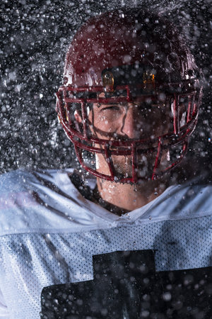 American Football Field: Lonely Athlete Warrior Standing on a Field Holds his Helmet and Ready to Play. Player Preparing to Run, Attack and Score Touchdown. Rainy Night with Dramatic Fog, Blue Lightの写真素材
