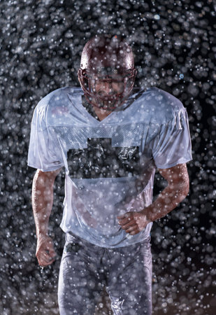 American Football Field: Lonely Athlete Warrior Standing on a Field Holds his Helmet and Ready to Play. Player Preparing to Run, Attack and Score Touchdown. Rainy Night with Dramatic Fog, Blue Lightの写真素材