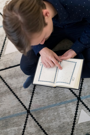Young muslim man sitting on the floor while reading holy book Quran before iftar dinner during a...