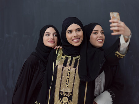 Group of young muslim women in fashionable dress with hijab using smartphone while taking selfie picture in front of black backgroundの写真素材