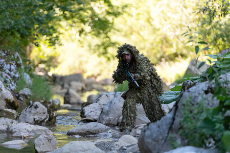 A military man or airsoft player in a camouflage suit sneaking the river and aims from a sniper rifle to the side or to target.の写真素材