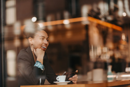 woman in a coffee shop drink coffee viewed through glass with reflections as they sit at a table chatting and laughingの写真素材