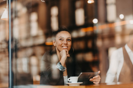 woman in a coffee shop drink coffee viewed through glass with reflections as they sit at a table chatting and laughingの写真素材