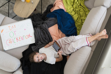 Top view of Little school girl with chickenpox playing creative games on tablet and resting on sofa at home, antiseptic cream applied to face and bodyの写真素材