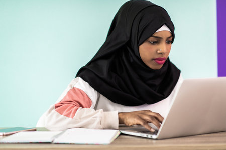afro girl wearing a hijab is disappointed and sad sitting in her home office and using a laptopの写真素材