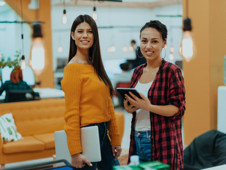 Portrait of two businesswomen talking to each other while standing in a modern business office with their colleagues using laptop and tablet.. Marketing concept. Multi-ethnic society.の写真素材