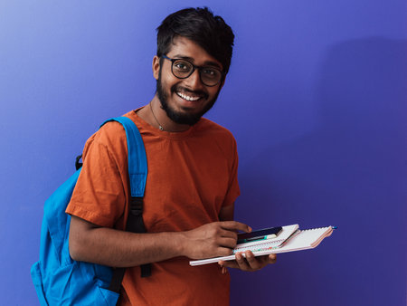 Indian student with blue backpack, glasses and notebook posing on purple background. The concept of...