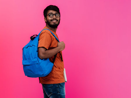 Indian student with blue backpack, glasses and notebook posing on pink background. The concept of...